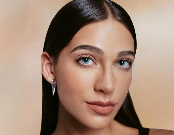 Rosto de mulher branca com maquiagem natural: pele lisa, sobrancelhas definidas, cílios longos, lábios rosados; cabelo castanho-escuro liso penteado para trás. Fundo bege suave e uniforme.