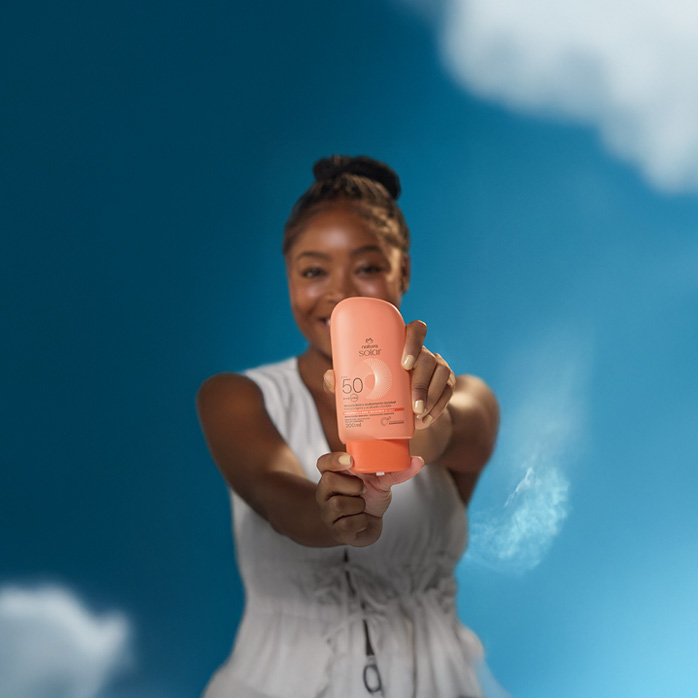 Mulher negra com cabelos trançados e presos segura à frente do corpo embalagem laranja do Protetor Solar Corporal FPS 50 Natura. Ela veste branco e sorri. O fundo é um céu azul, com poucas nuvens.