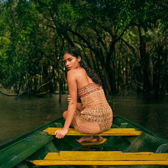 Uma mulher morena em um barco de madeira, cercada por águas calmas e vegetação densa, transmite conexão plena com a Amazônia, refletindo o espírito livre e autêntico da linha Natura Ekos Ryos.