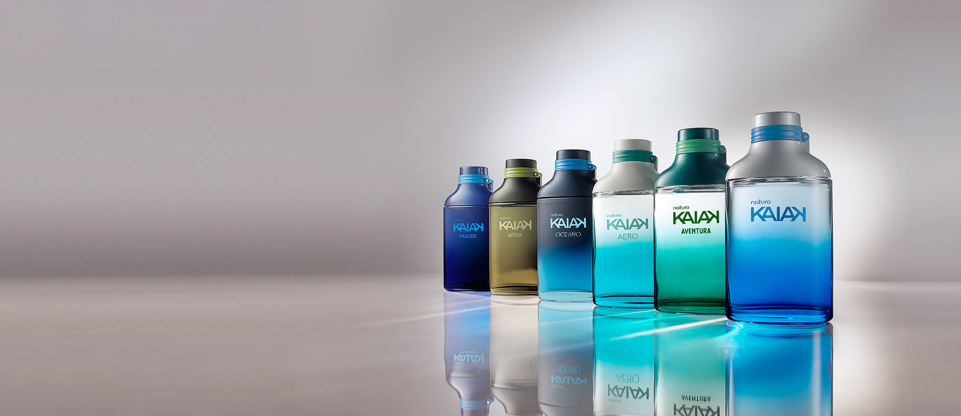 Frascos de Natura Kaiak: Pulso em azul-escuro, Urbe verde-escuro, Oceano em tons de azul, Aero em azul-claro, Aventura em verde e Kaiak em azul sobre superfície reflexiva com fundo cinza-claro.