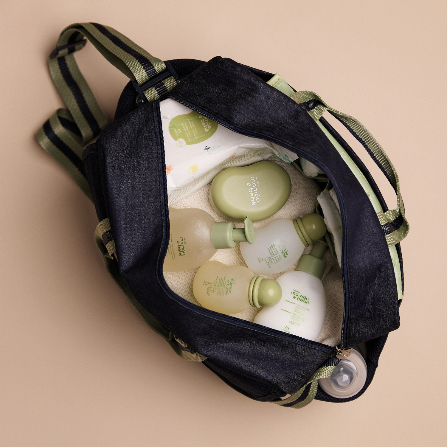 Bolsa Mamãe e Bebê aberta sobre superfície bege. Dentro da bolsa é possível ver uma toalha, várias fraldas e produtos mamãe e bebê. A bolsa contém bolso lateral onde há uma mamadeira. 