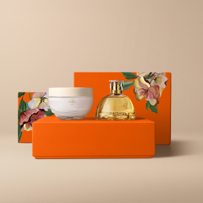 Presente Natura Aura Alba Completo, contendo um Deo Parfum e um Creme Aveludado Corporal. Os itens estão sobre caixas de presentes laranja com estampa de flores em fundo bege.
