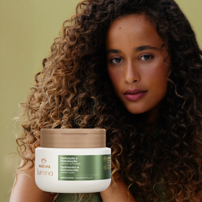 Pote branco com rótulo verde da Máscara Lumina Definição e Hidratação. Uma mulher negra com cabelos castanhos cacheados está em primeiro plano, sobre fundo verde.