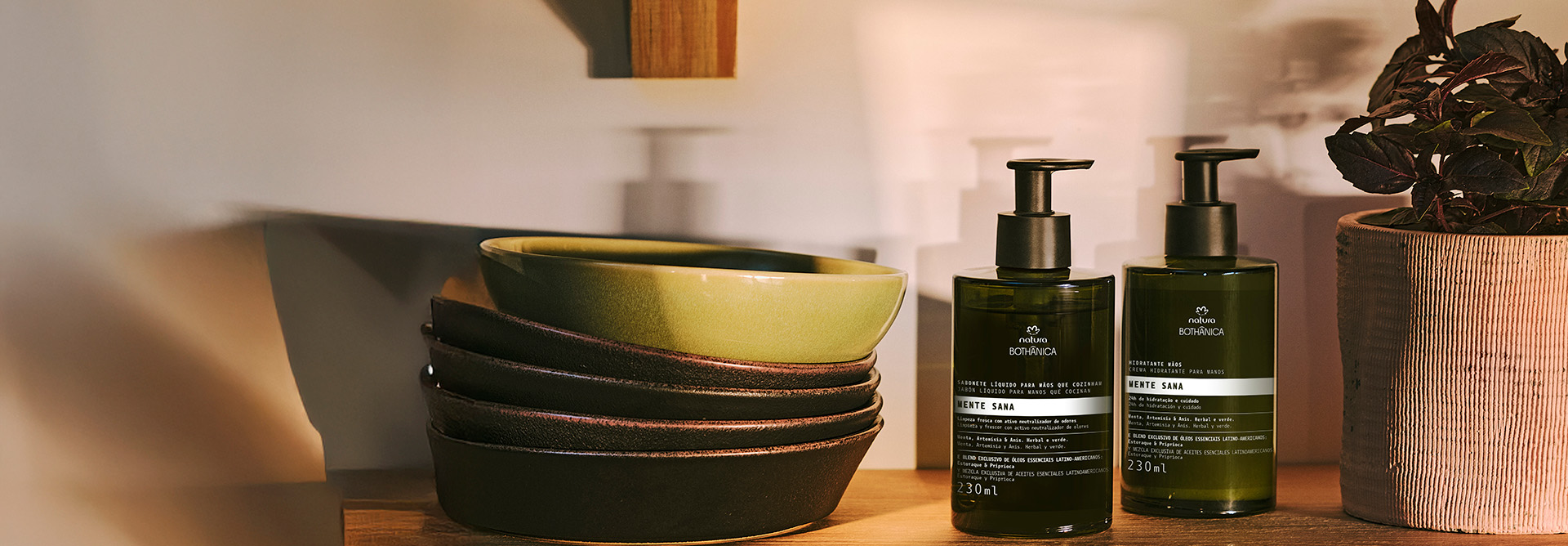 2 frascos verde-escuros de Bothânica Mente Sana. 1 Sabonete Líquido para Mãos que Cozinham e 1 Hidratante para Mãos. Os produtos estão sobre uma prateleira, com tigelas de cerâmica e plantas ao lado.