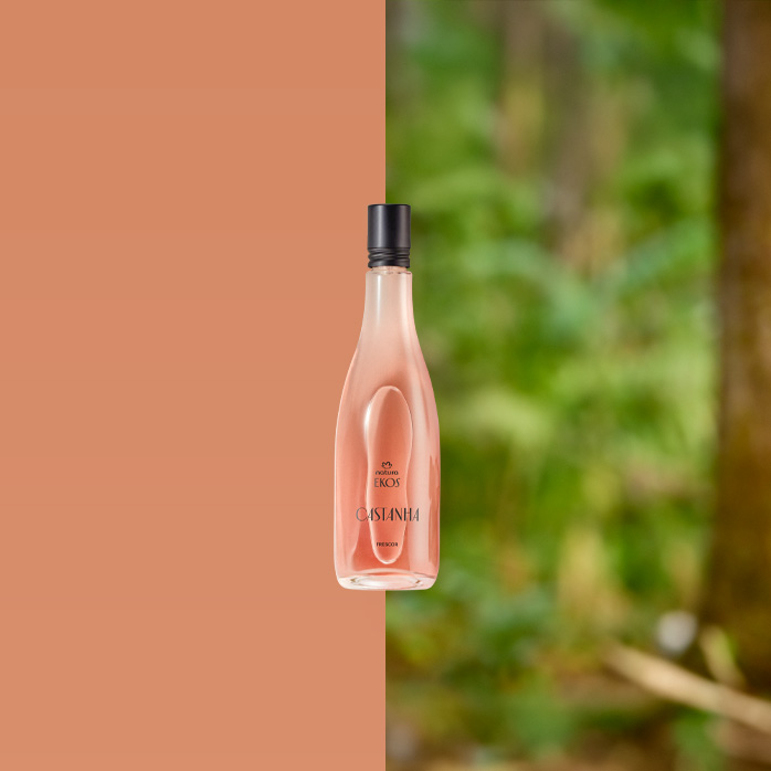 Frasco do Deo Parfum Ekos Castanha em tom rosado com tampa preta está apoiado sobre um tronco. Ao fundo, folhas verdes e galhos compõem um cenário de floresta iluminada.