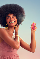 Modelo negra de cabelos crespos usa vestido longo rosê com brincos e bracelete dourados. Ela sorri para a câmera e borrifa o Perfume para Cabelos Luna Divina.