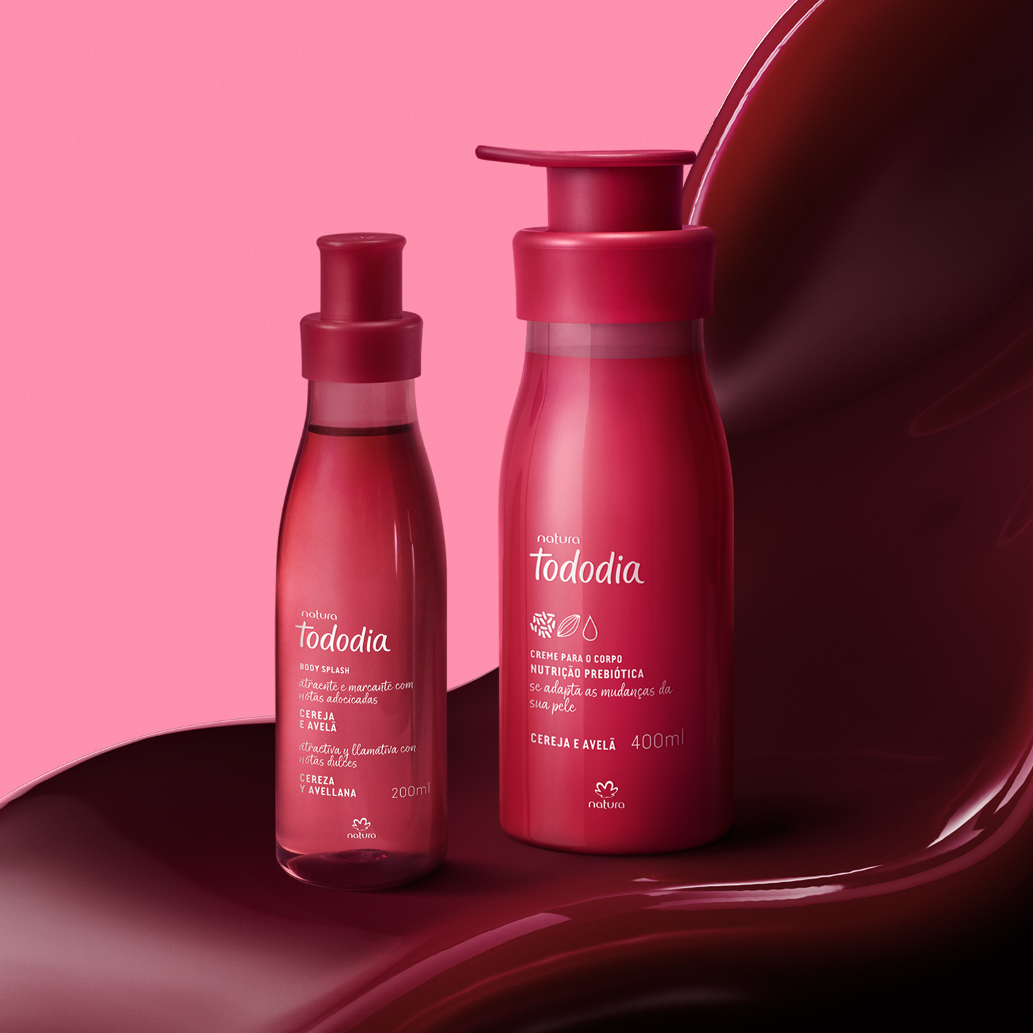 2 produtos de Tododia Cereja e Avelã, sendo 1 frasco de body splash e 1 frasco pump de creme, ambos com embalagens vermelhas, repousam sobre uma textura cremosa na cor cereja.