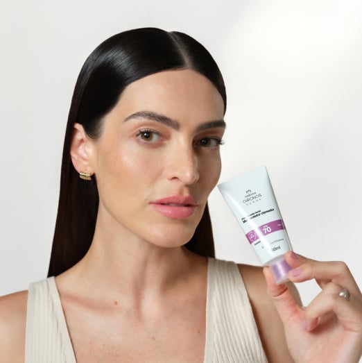 Mulher negra segura Multiprotetor FPS 70 próximo ao rosto. Ela usa blusa branca de ombro único e está em fundo branco.