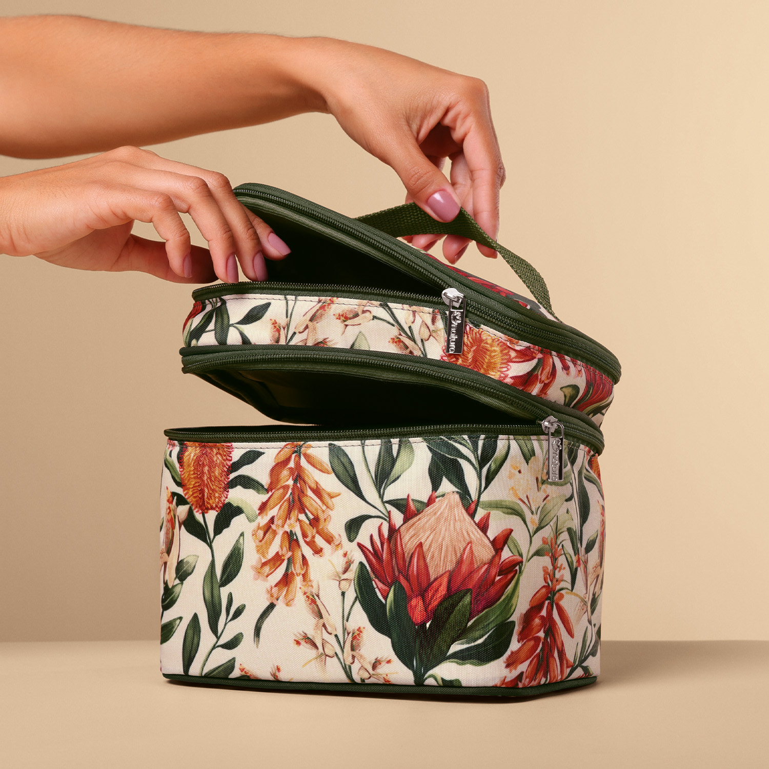 Mãos femininas abrindo Frasqueira Natura em fundo neutro. Estampa floral delicada em tons de verde, laranja e vermelho.