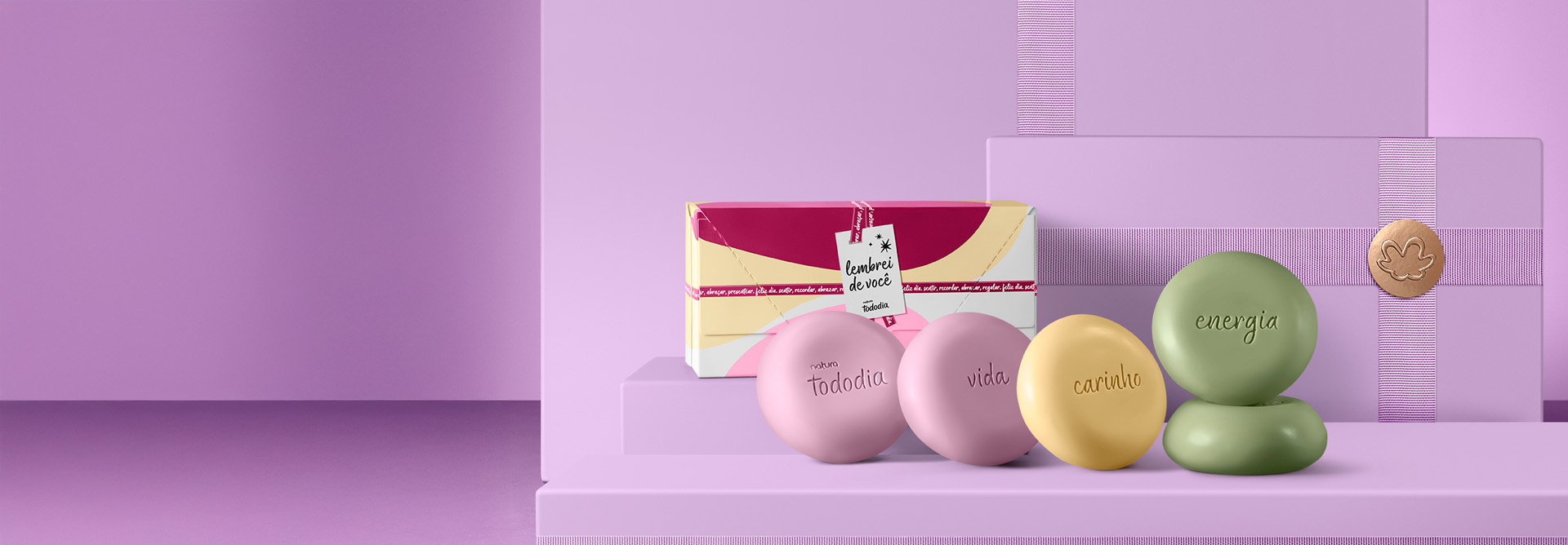 Presente Tododia com caixa e sabonetes nas cores rosa, amarelo e verde com as inscrições Natura Tododia, vida, carinho e energia, sobre caixas presenteáveis lilás com selo dourado.