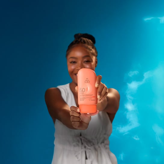 Mulher negra de cabelos trançados presos, vestindo branco, exibe embalagem laranja de Natura Solar Corporal FPS 50, em fundo de céu azul.