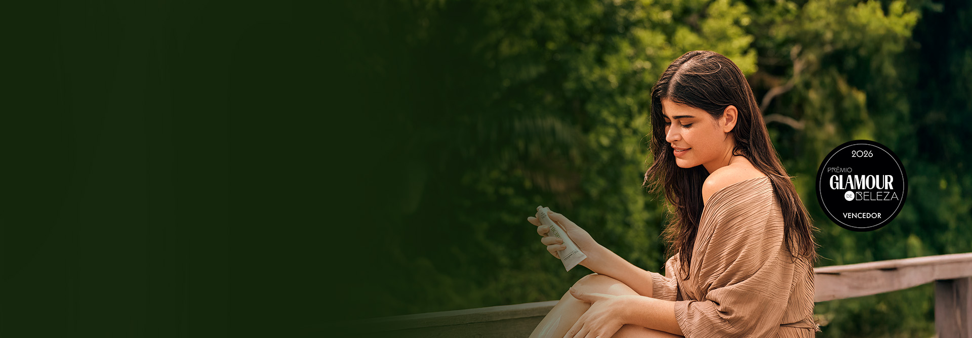 Uma mulher de cabelos castanhos ondulados e pele clara, vestindo um roupão bege, aplica creme nas pernas. Ela está sentada ao ar livre com um fundo desfocado de vegetação verde exuberante. Ela sorri suavemente enquanto segura uma bisnaga de hidratante prateada. Selo circular preto do "Prêmio Glamour de Beleza 2026", com a palavra "Vencedor" na parte inferior.