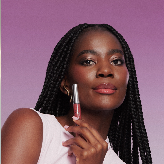 Modelo negra de cabelos longos aparece no centro da tela, em fundo roxo, com o Gloss Labial Natura Una nos lábios. Ela segura o Gloss Labial Natura Una próximo ao rosto.