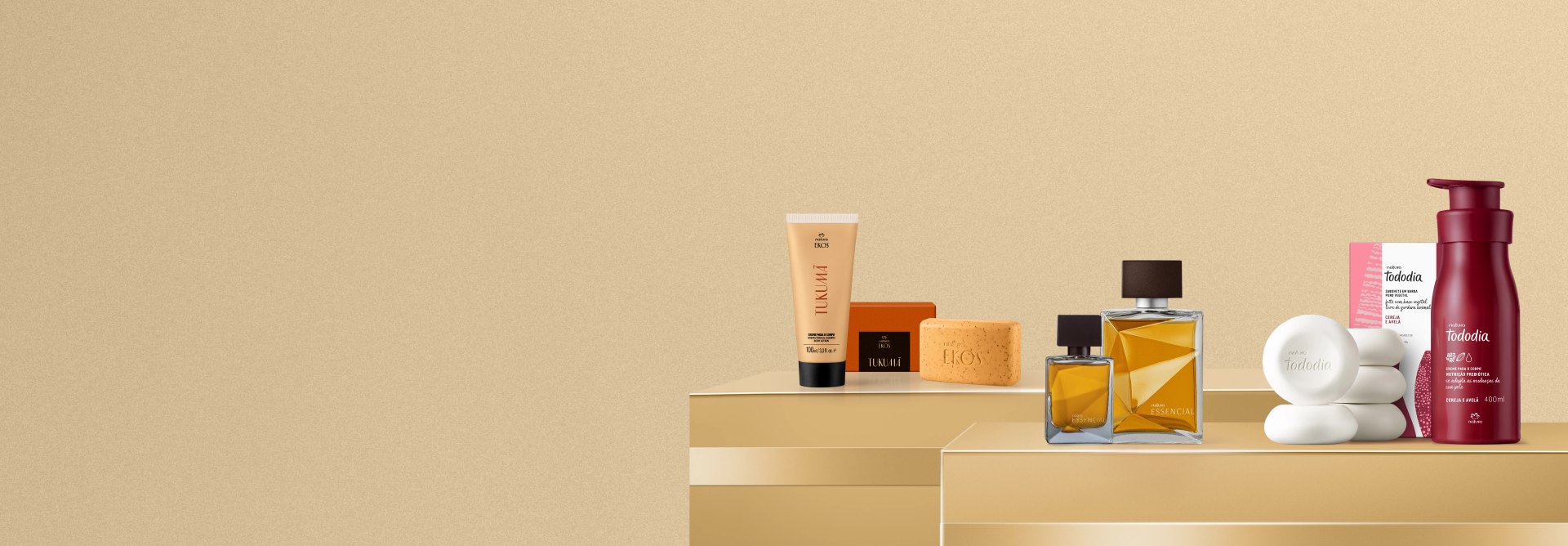 Produtos das linhas Ekos Tukumã, Essencial Masculino e Tododia Cereja e Avelã dispostos sobre prateleiras douradas com laços na mesma cor. Fundo liso bege.