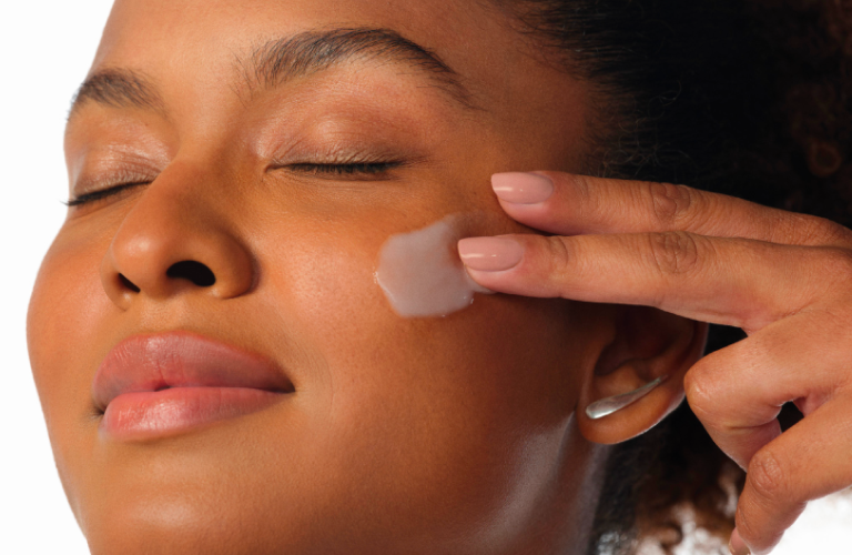 Mulher aplicando um creme Chronos Derma próximo ao rosto, com a embalagem branca encostada na bochecha.