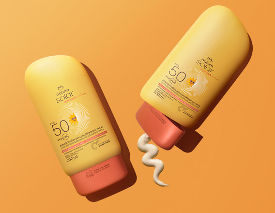Duas embalagens de protetor solar Natura Solar FPS 50 infantil. Ambas são amarelas com detalhes em laranja avermelhado. Uma das embalagens está deitada na horizontal, com a tampa aberta e o protetor solar branco cremoso espremido para fora em um formato ondulado. A outra embalagem está posicionada verticalmente ao lado dela. O fundo é de um tom laranja vibrante.
