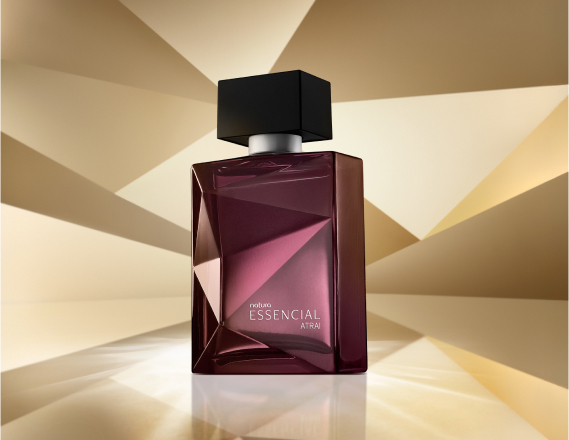 Frasco de Essencial Atrai Masculino, em tons de roxo e violeta com tampa preta, aparece em destaque em fundo dourado multifacetado, fazendo alusão ao design dos frascos de Essencial.