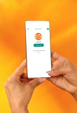 Consultora de Beleza Natura e Avon, de pele morena e cabelos escuros e curtos, está sentada à mesa escrevendo. À sua frente, revistas Natura e Avon, app Natura no celular e site Natura no notebook.