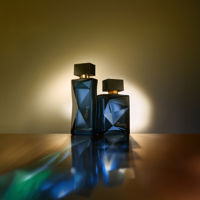 Essencial Oud Feminino e Masculino, com frascos em tons de azul-escuro e tampas pretas, iluminam a superfície âmbar em que estão apoiados com seus reflexos azul-esverdeados em fundo dourado.