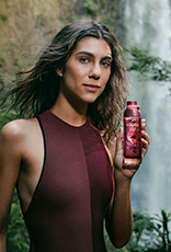 Modelo morena de cabelos lisos e soltos usa maiô vinho e segura o novo Kaiak Aventura Intensa Feminino nas mãos. Ao fundo, destaca-se a imagem de uma cachoeira em meio à floresta.