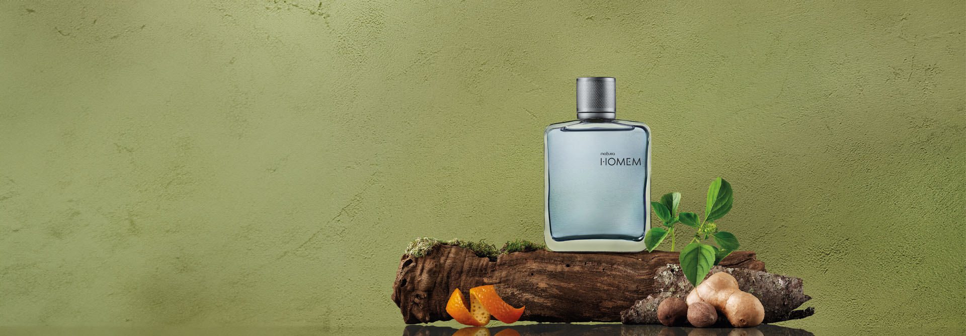 Desodorante Colônia Natura Homem disposto à direita sobre um pedaço de madeira e outros ingredientes que compõem a fragrância.