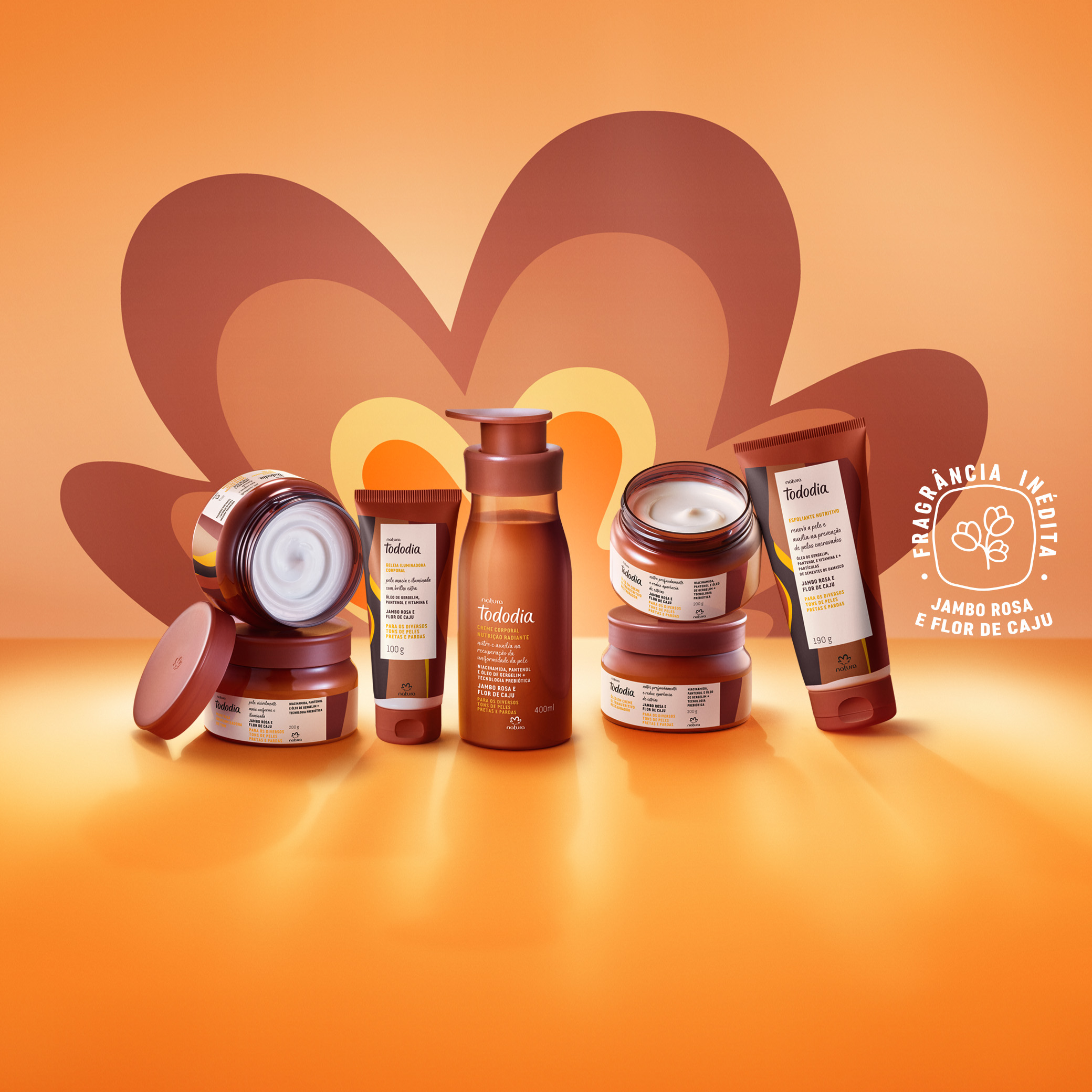 7 produtos Tododia na fragrância Jambo Rosa e Flor de Caju, em potes, bisnagas e frascos. Fundo em tons de laranja e marrom. 