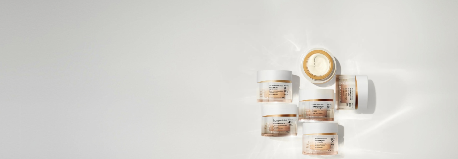 Cremes Antissinais Chronos Derma em fundo branco iluminado. Seis potes translúcidos com degradê branco, tampa branca e detalhes em dourado. Um deles está aberto e mostra conteúdo cremoso bege dentro.