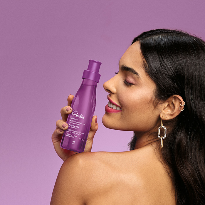 Mulher de cabelos soltos está de costas e segurando sobre o ombro esquerdo um Body Splash Natura  Tododia em fundo lilás