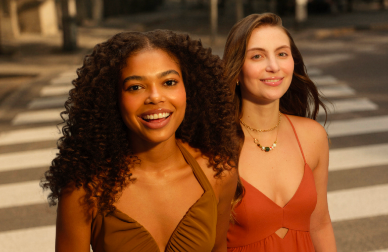 Duas jovens sorridentes caminhando por uma faixa de pedestre, uma com vestido marrom e cabelo cacheado solto, a outra com vestido terracota e colar dourado.