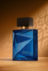 Deo Parfum Essencial Oud Masculino em frasco facetado azul com tampa preta. Está em fundo cor areia com sombra geométrica projetada.