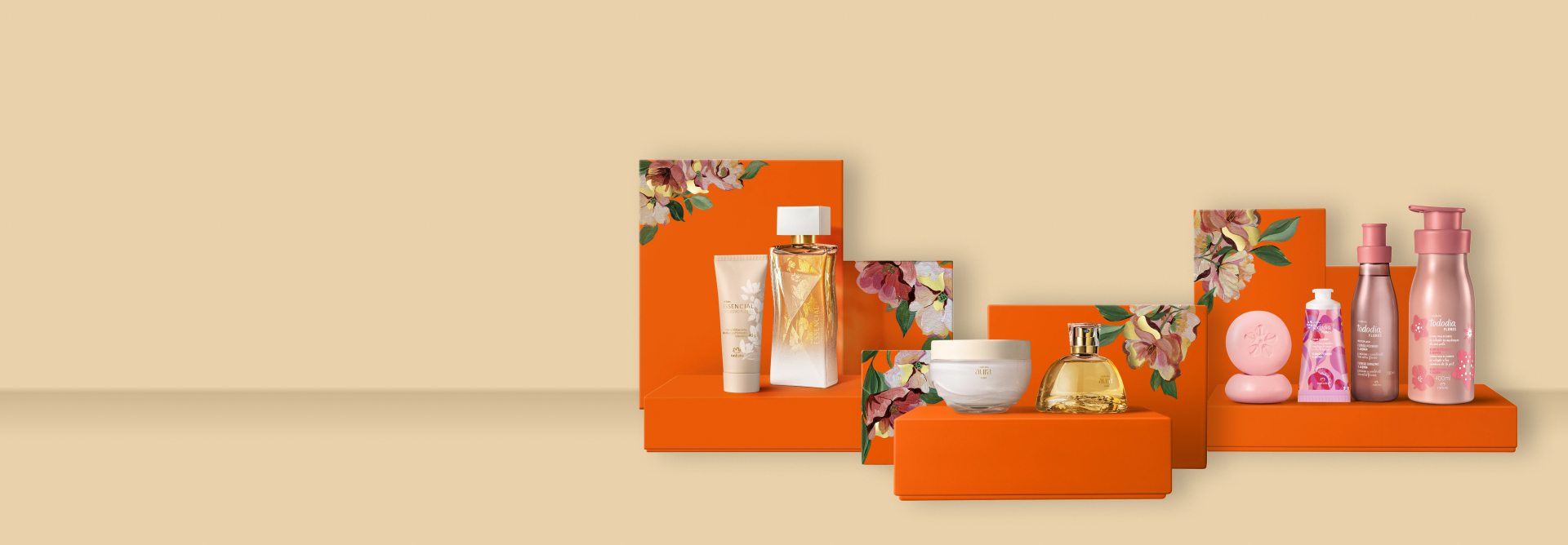 Presente Natura Essencial, Presente Natura Aura Alba e Presente Natura Flores Essenciais sobre caixas de presentes laranja com estampa floral em fundo bege.