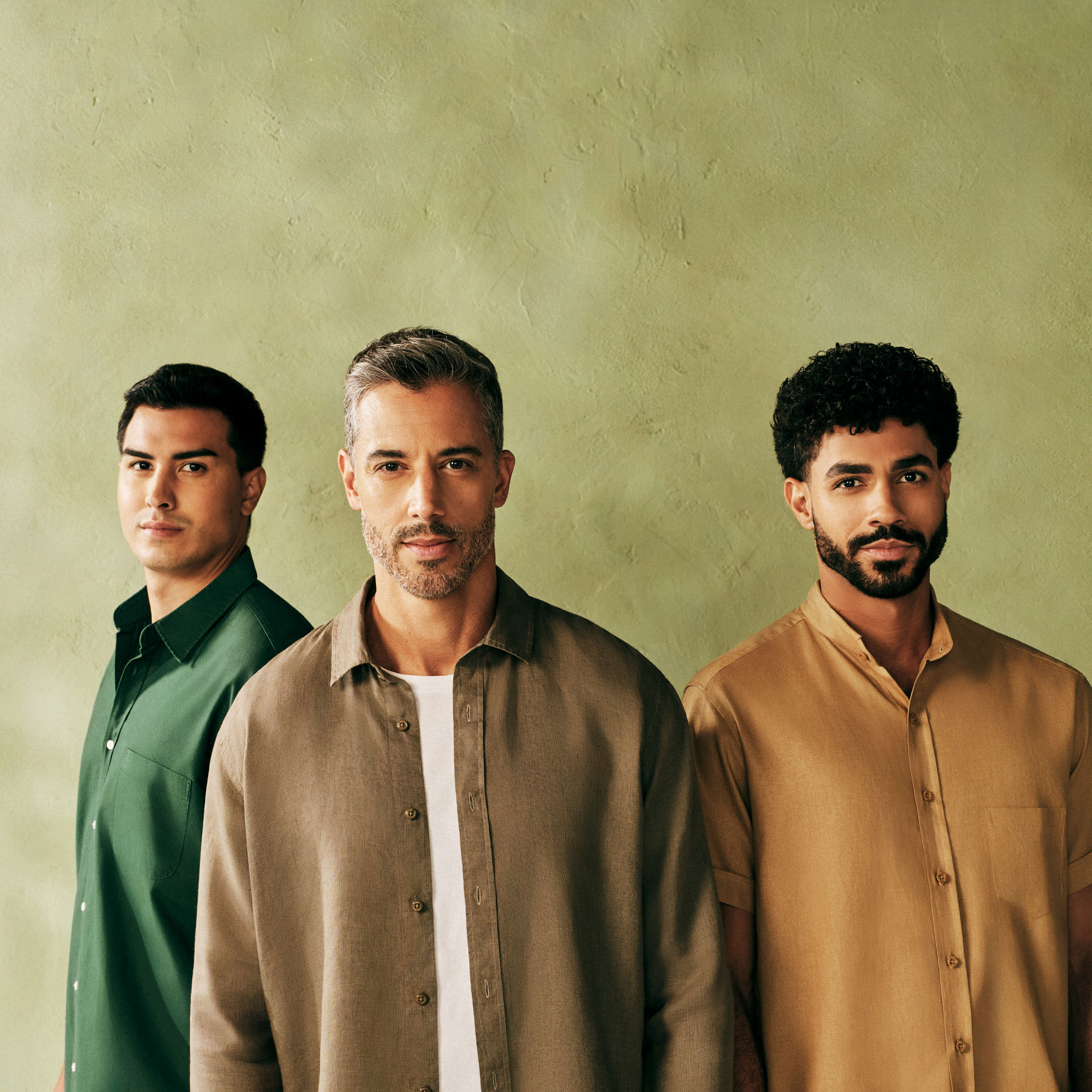 Foto de três modelos homens olhando fixamente para a câmera, o primeiro está de perfil e os demais, de frente. Todos têm cabelos curtos e usam camisas. O fundo da imagem é verde.