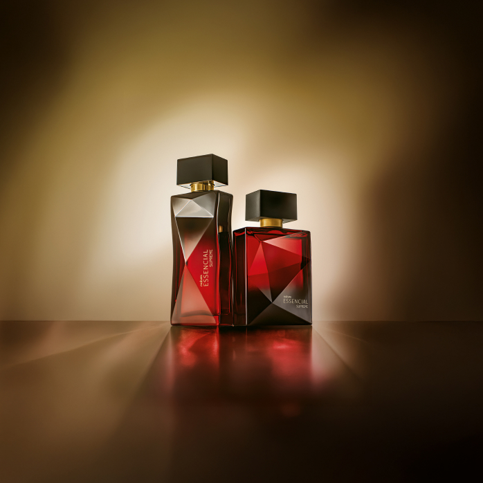 Essencial Supreme Feminino e Masculino, com frascos em tons de preto e vermelho, aparecem iluminados sobre superfície marrom e fundo dourado.