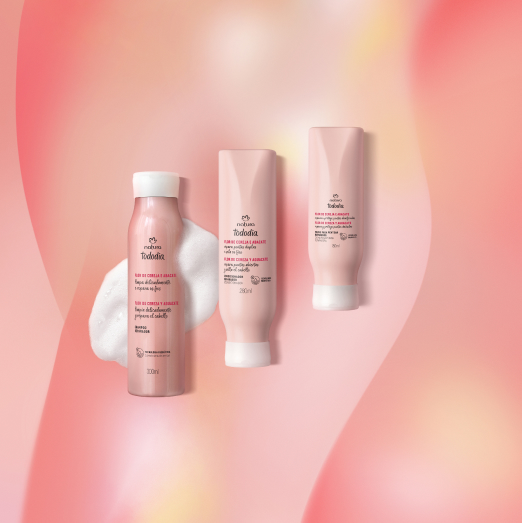 Três produtos Natura Tododia Flor de Cereja e Abacate para cabelos. Um shampoo com espuma, um condicionador e um creme de pentear em embalagens rosa-claro. Fundo com degradê de rosa.