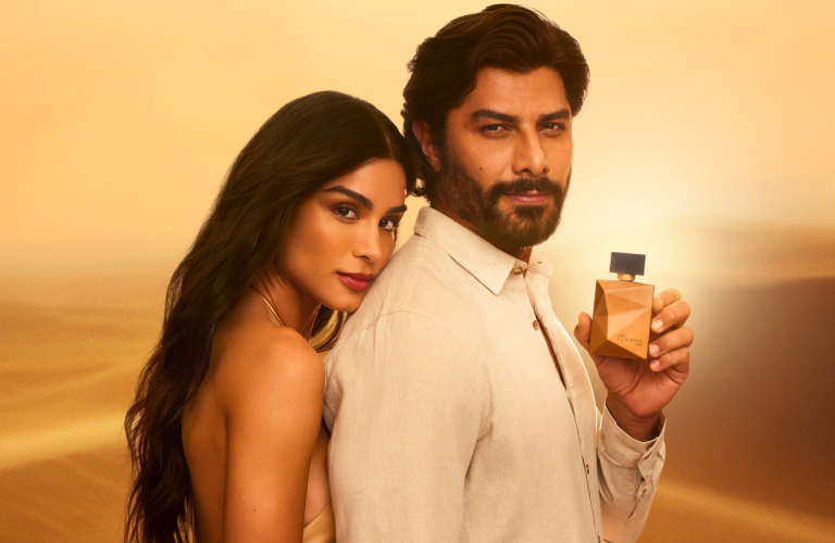Foto com fundo desértico e dourado exibindo um homem de camisa clara segurando um perfume Natura Essencial, ao lado de uma mulher de cabelos longos inclinada em seu ombro.