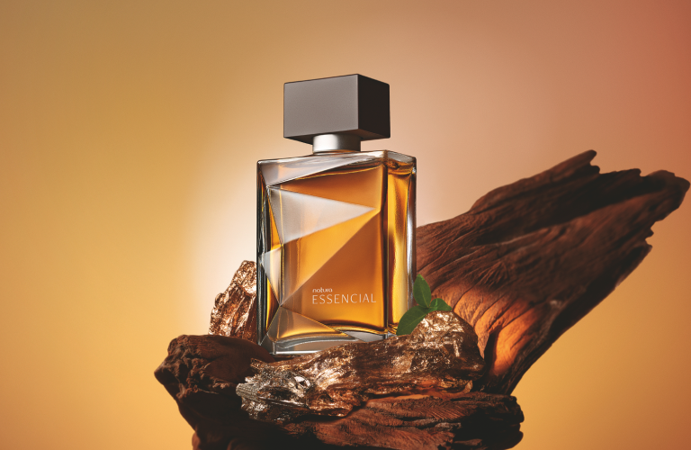 Foto com fundo degradê em tons quentes de âmbar e dourado destacando um frasco de perfume Natura Essencial apoiado sobre um pedaço de madeira rústica com detalhes iluminados.