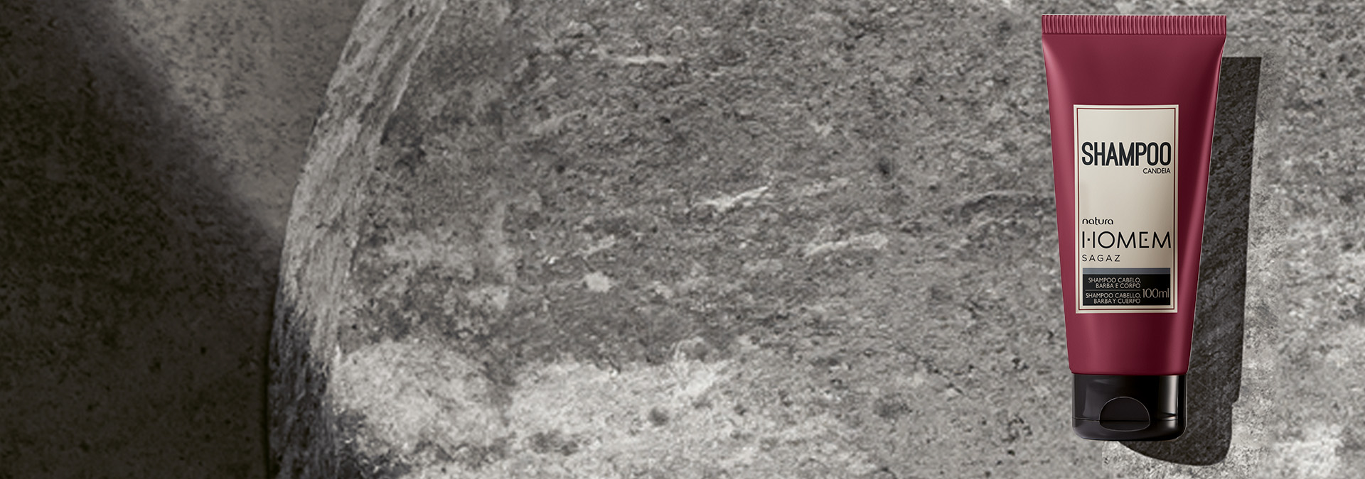 Fondo gris oscuro con una rosácea en el lado derecho, y frente a la rosácea, dos champús de la línea Homem de Natura.