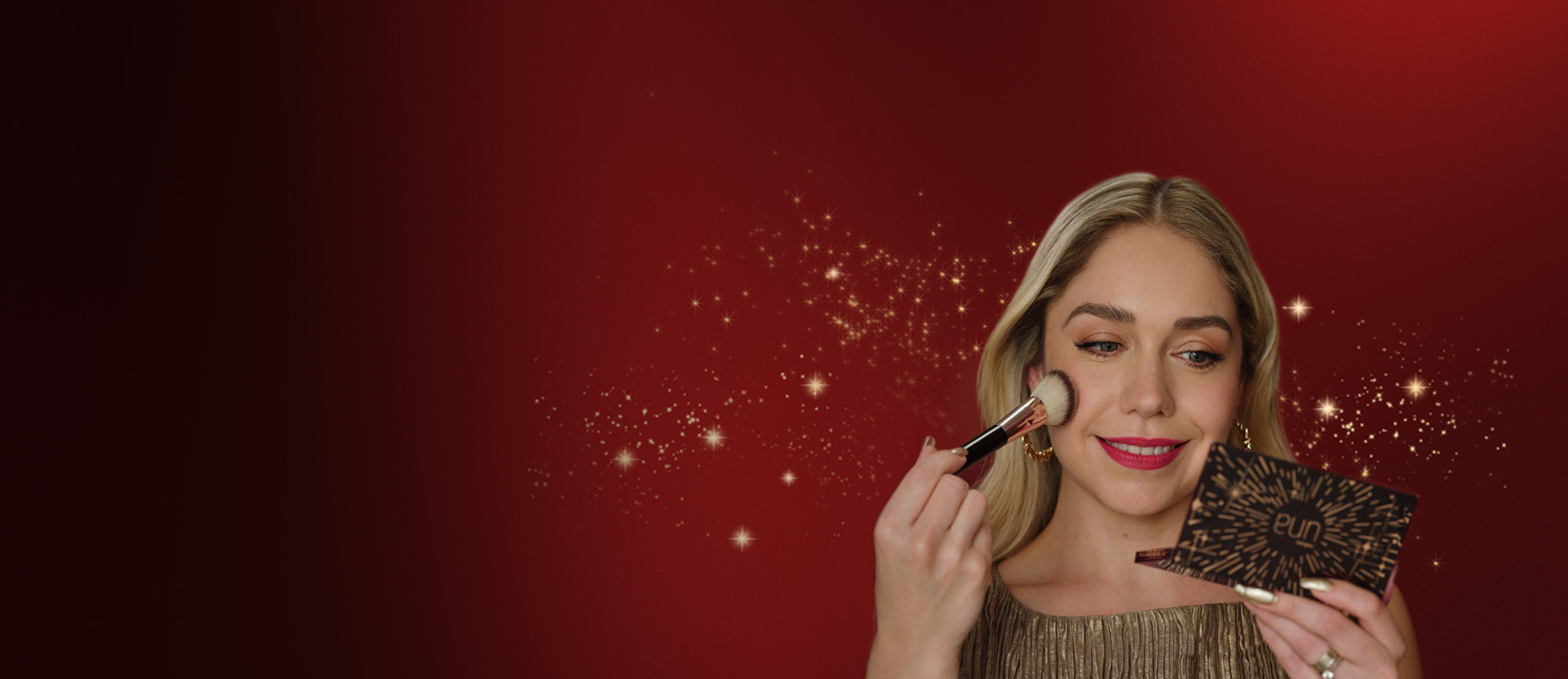 Fondo rojo con brillos navideños, y en primer plano, una mujer blanca y rubia aplicando maquillaje con un pincel de una paleta de maquillaje de Natura.