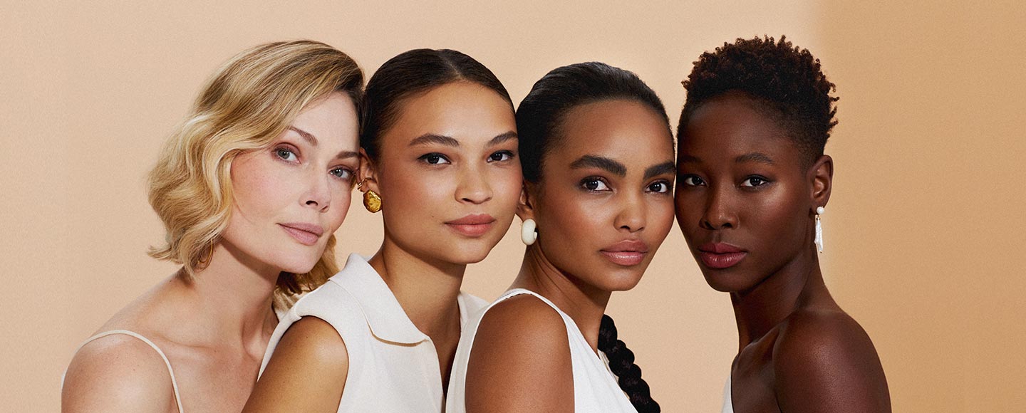 Cuatro mujeres de diferentes etnias y edades posan juntas, hombros cercanos, con un fondo beige imitando polvo compacto.