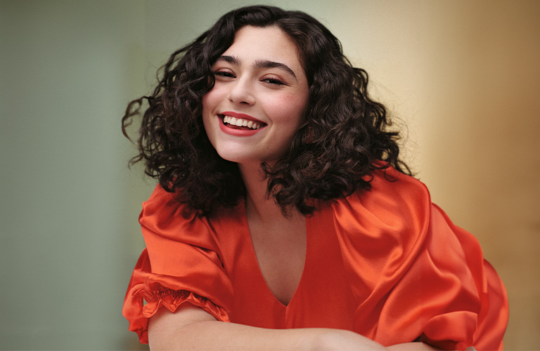 Foto de una mujer con cabello corto y rizado. El fondo tiene tonos verdes y amarillos, y lleva una blusa roja con mangas abullonadas.