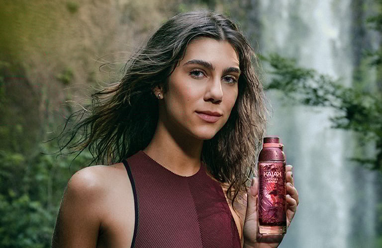 Una mujer con traje de baño rojo sostiene un frasco de perfume Kaiak rojo. Al fondo, una cascada y vegetación.