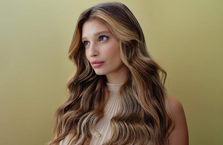 Foto de una mujer con cabello ondulado, largo y con mechas en tonos castaños y rubios. El fondo es amarillo claro, y lleva una blusa beige con textura delicada.