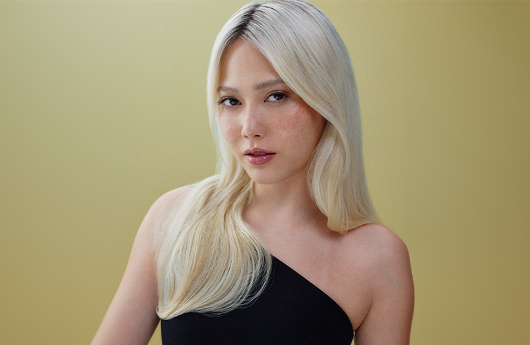 Foto de una mujer con cabello liso, largo y rubio, peinado hacia un lado. El fondo es amarillo claro, y lleva una blusa negra de un solo hombro.
