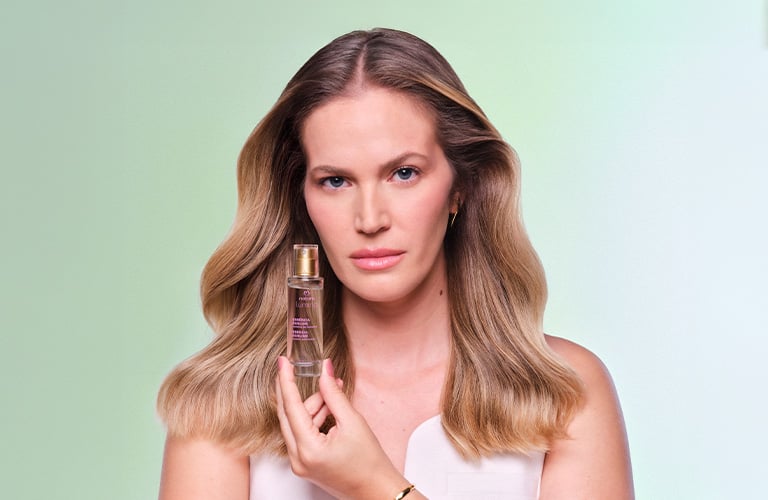 Mujer con cabello rubio y ondulado sosteniendo un frasco de perfume capilar. Fondo verde claro.