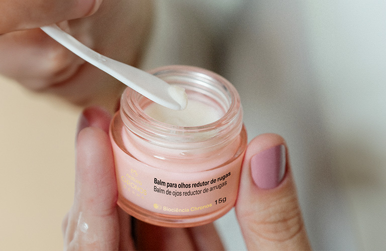 Fotografía de unas manos sosteniendo un tarro de bálsamo para ojos, con fondo neutro. Una espátula blanca está dentro del tarro, que es de color rosa.