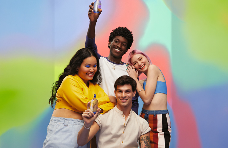 Foto con fondo de colores degradados, cuatro personas jóvenes posan en grupo, dos sostienen frascos de perfume unisex hacia la cámara.