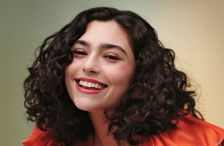 Foto de una mujer con cabello corto y rizado. El fondo tiene tonos verdes y amarillos, y lleva una blusa roja con mangas abullonadas.