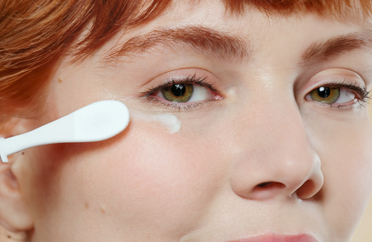 Fotografía de una mujer con el cabello rojizo aplicando crema blanca bajo el ojo, sobre un fondo neutro.