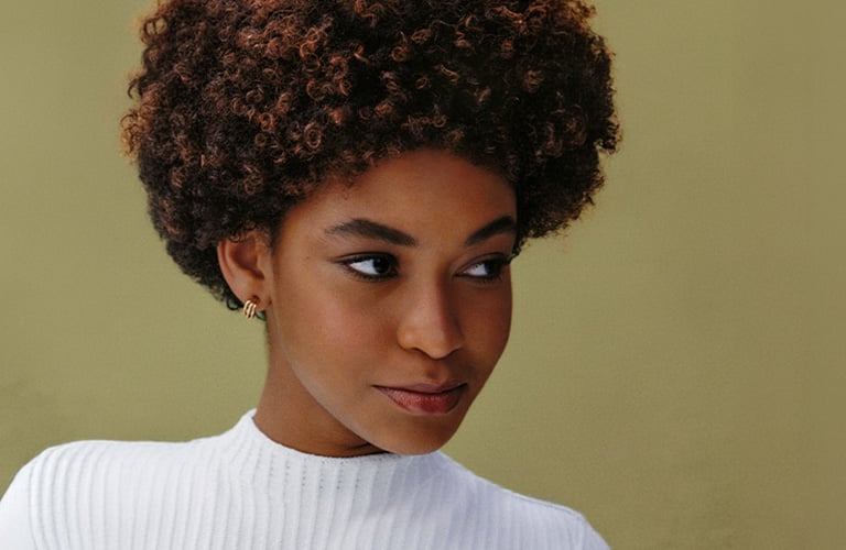 Foto de una mujer con cabello crespo corto en tonos castaños. El fondo es verde, y lleva una blusa blanca sin mangas mientras sostiene un mechón de su cabello con la mano izquierda.