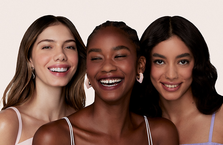 Foto de tres mujeres con diferentes tipos de cabello. El fondo es beige claro, y llevan ropa en tonos pastel: rosa, blanco y lila.